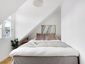 A bedroom with a sloped ceiling, a bed with a marble headboard, a bedside table with a lamp, and a window with a view of buildings. - Saint - Nice 1-bdr Apt by the Canals (Copenhagen)