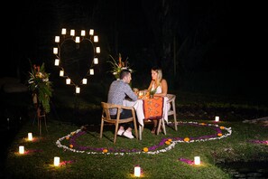 Um casal jantando ao ar livre à noite, com uma decoração de luz em formato de coração e arranjos florais na grama.