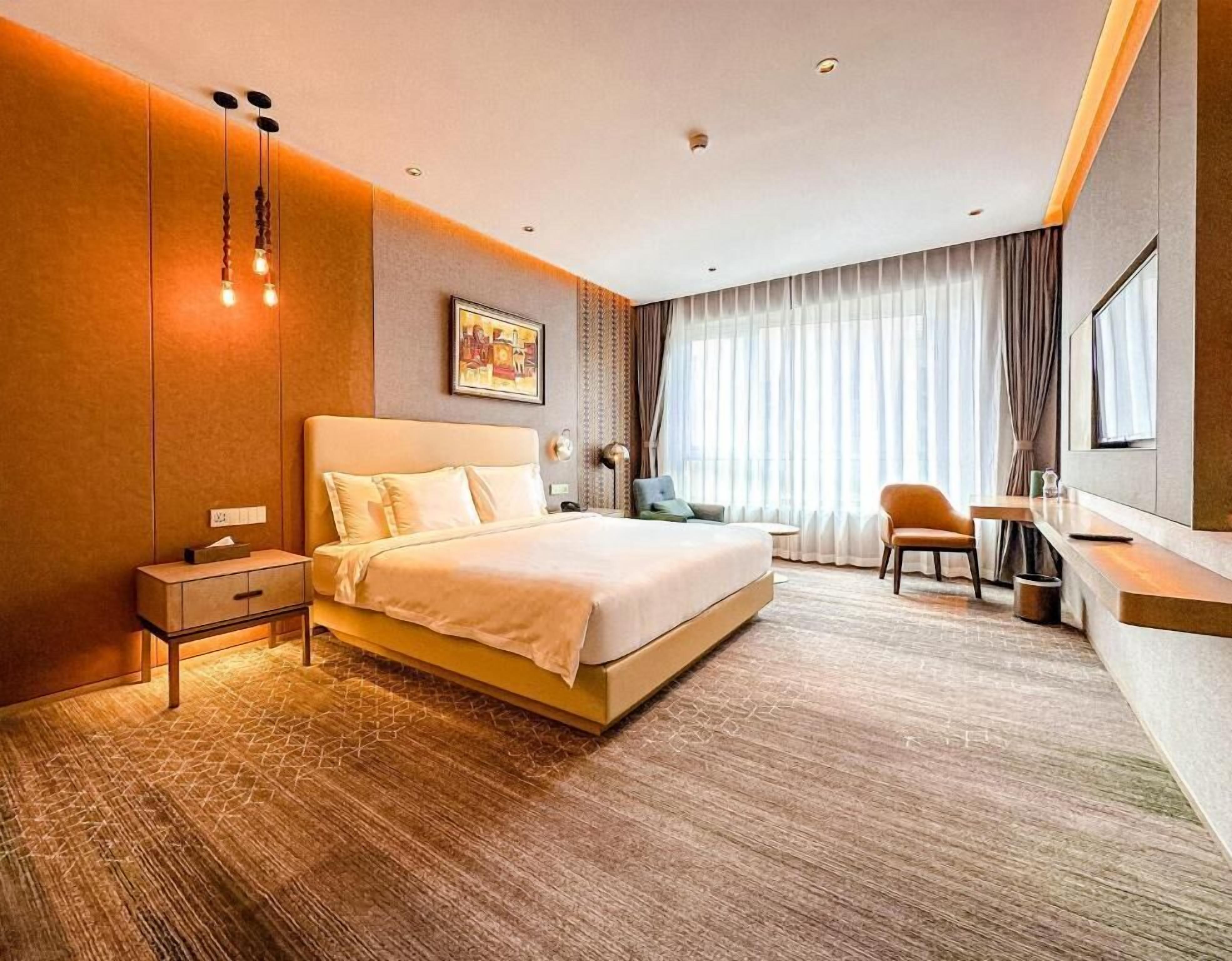 A hotel room with a large bed, bedside table, lamp, chair, and a view of the city through the window.