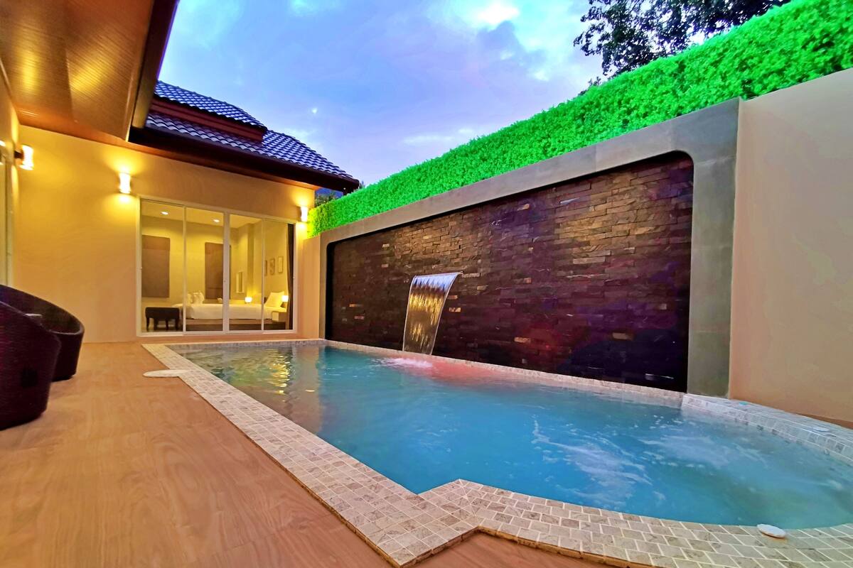 A modern indoor swimming pool with a waterfall feature, surrounded by a wooden deck and a glass door leading to a room with a bed and a sofa.