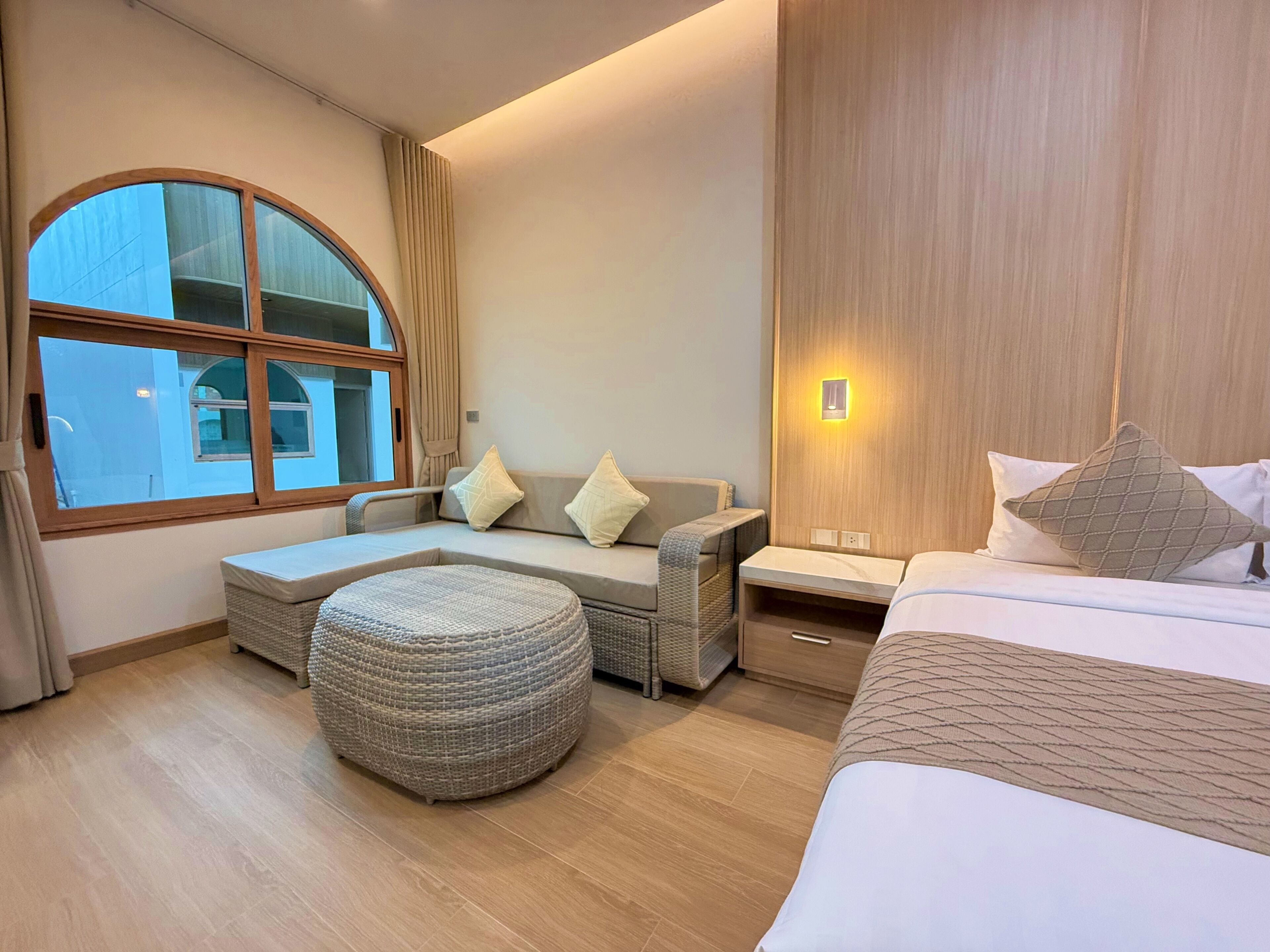 A hotel room with a large window, a sofa with cushions, a small ottoman, a bedside table, and a bed with pillows.