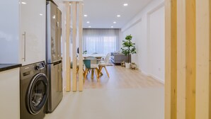 A modern kitchen with a built-in washer and dryer, a dining area with wooden chairs, and a large window with sheer curtains.