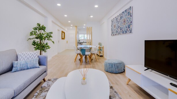 A modern living room with a grey sofa, a white coffee table, a TV stand, and a dining area with a table and chairs.