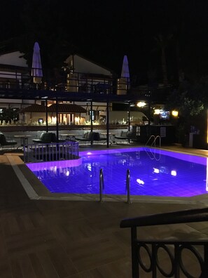 Zona de piscina nocturna amb una piscina iluminada de blau, mobiliari d'exterior i una zona de dinar coberta.