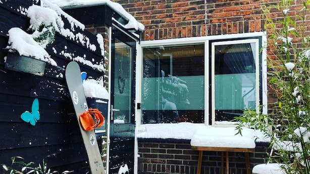 A snow-covered outdoor area with a snowboard leaning against a wall, a window, and a wooden table.