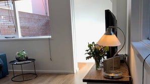 A modern living room with a wooden floor, a round table with a plant, a lamp, and a TV on a stand.