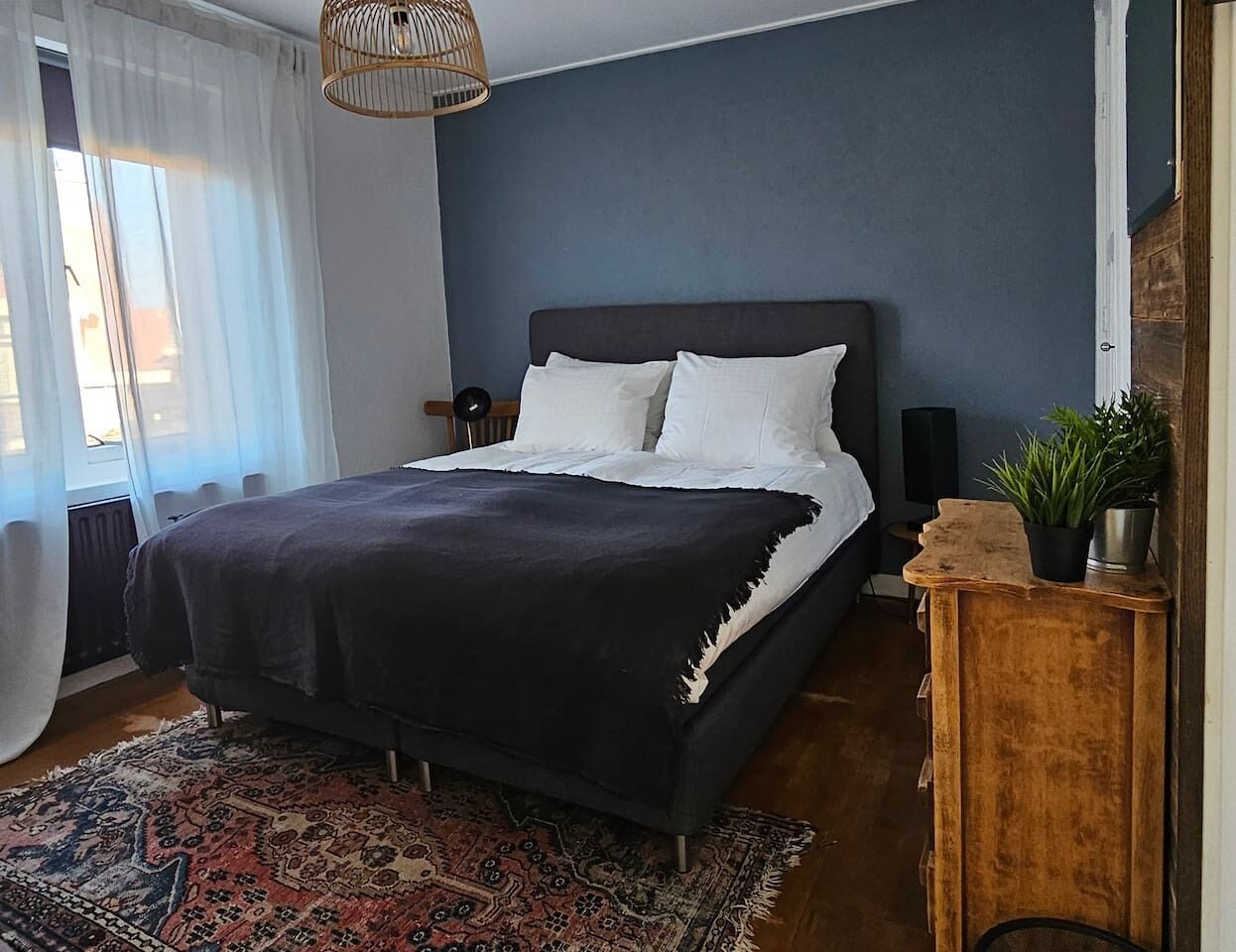 Quarto com cama, mesa de cabeceira, luminária, planta em vaso e uma grande janela com cortinas.