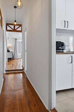 A hallway with wooden flooring leading to a kitchen area with white cabinets and a coffee machine.