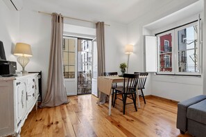 A room with a wooden floor, a dining table with chairs, a TV on a cabinet, and a window with a view of buildings.