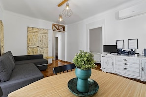 A modern living room with a grey sofa, a wooden dining table with a green plant centerpiece, a white cabinet with audio equipment, and a wooden-framed wall decoration.