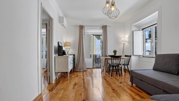 Una sala de estar moderna con sofá, mesa de comedor y sillas. Hay un televisor sobre un mueble bajo, una lámpara y una ventana con cortinas.