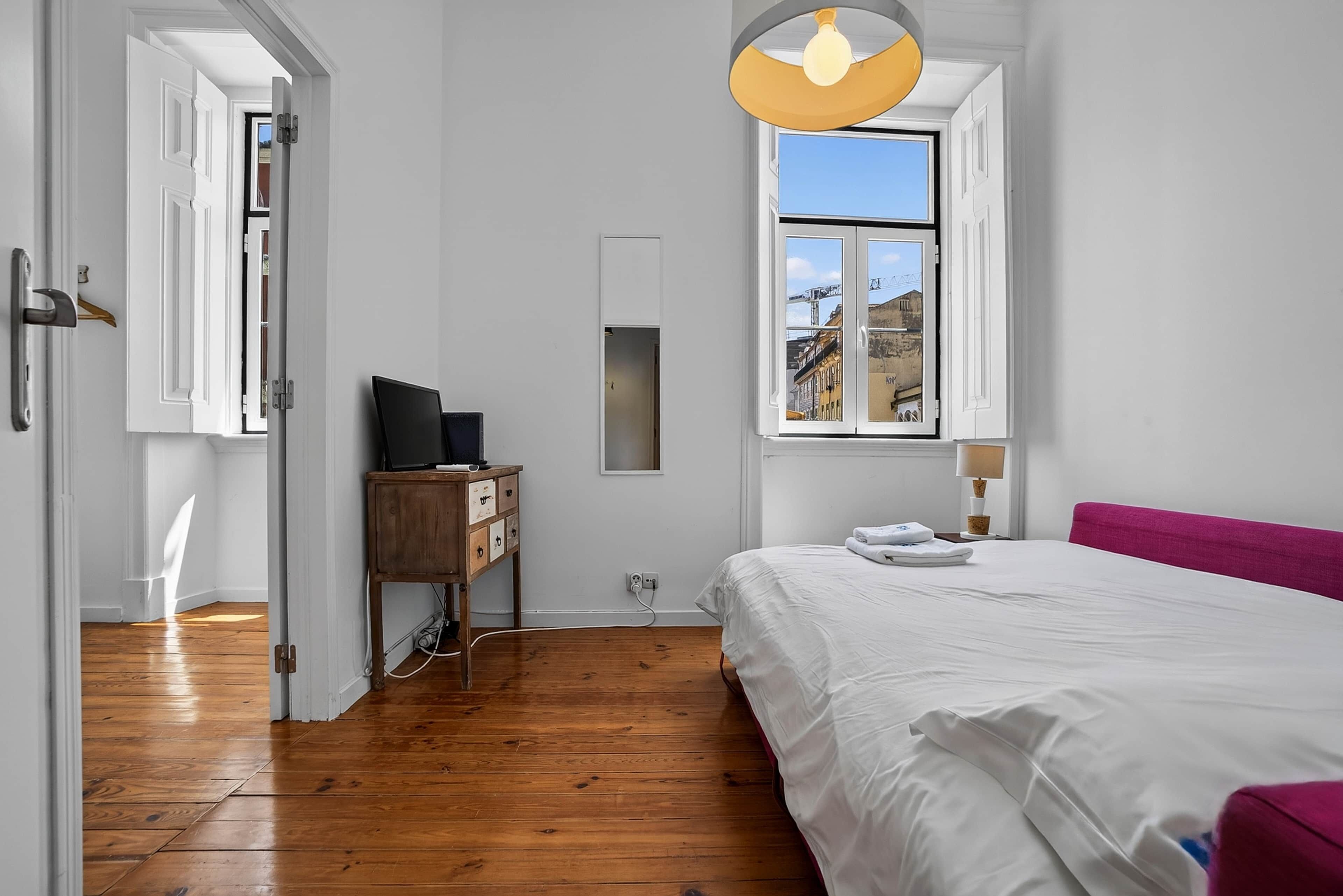 Een slaapkamer met houten vloer, een bed met wit beddengoed, een bureau met een computer, een raam met uitzicht op gebouwen, en een deur naar een andere kamer.