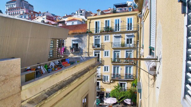 Une rue étroite, bordée de bâtiments des deux côtés, avec des balcons dotés de balustrades et des espaces de détente en plein air abrités par des parasols.