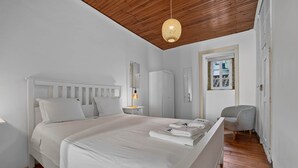 Un dormitorio con cama blanca, mesita de noche con lámpara, armario y una silla.