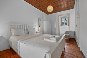 Ein Schlafzimmer mit einem weißen Bett, einem Nachttisch mit Lampe, einem Kleiderschrank und einem Sessel.