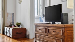 Un aparador de madera con un televisor encima, una maleta y una planta en una habitación con piso de madera y una ventana.