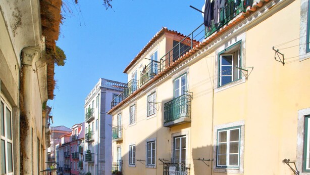 Una calle estrecha flanqueada por edificios, con mesas al aire libre y sombrillas, bajo un cielo azul despejado.