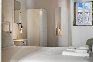 A hotel room with a bed, a nightstand, a lamp, a wardrobe, a mirror, and a window with a view of buildings.