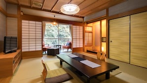 Une pièce de style japonais traditionnel, avec un sol recouvert de tatamis, une table basse, un téléviseur, des portes coulissantes et une vue sur la verdure à l’extérieur.
