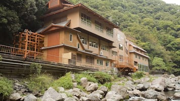 A traditional Japanese building with multiple floors, a wooden structure on the left, and a stone bridge over a rocky stream.