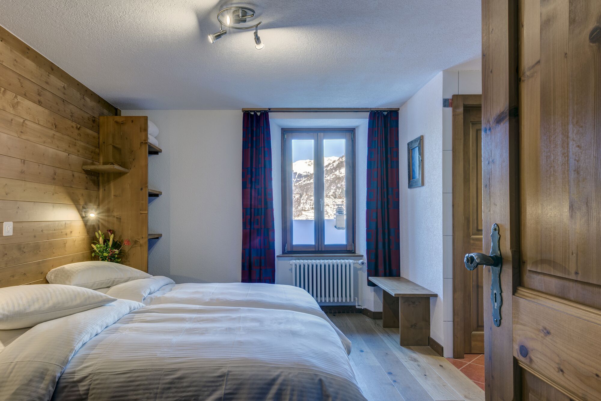 A bedroom with two beds, a window with curtains, a radiator, and wooden walls.