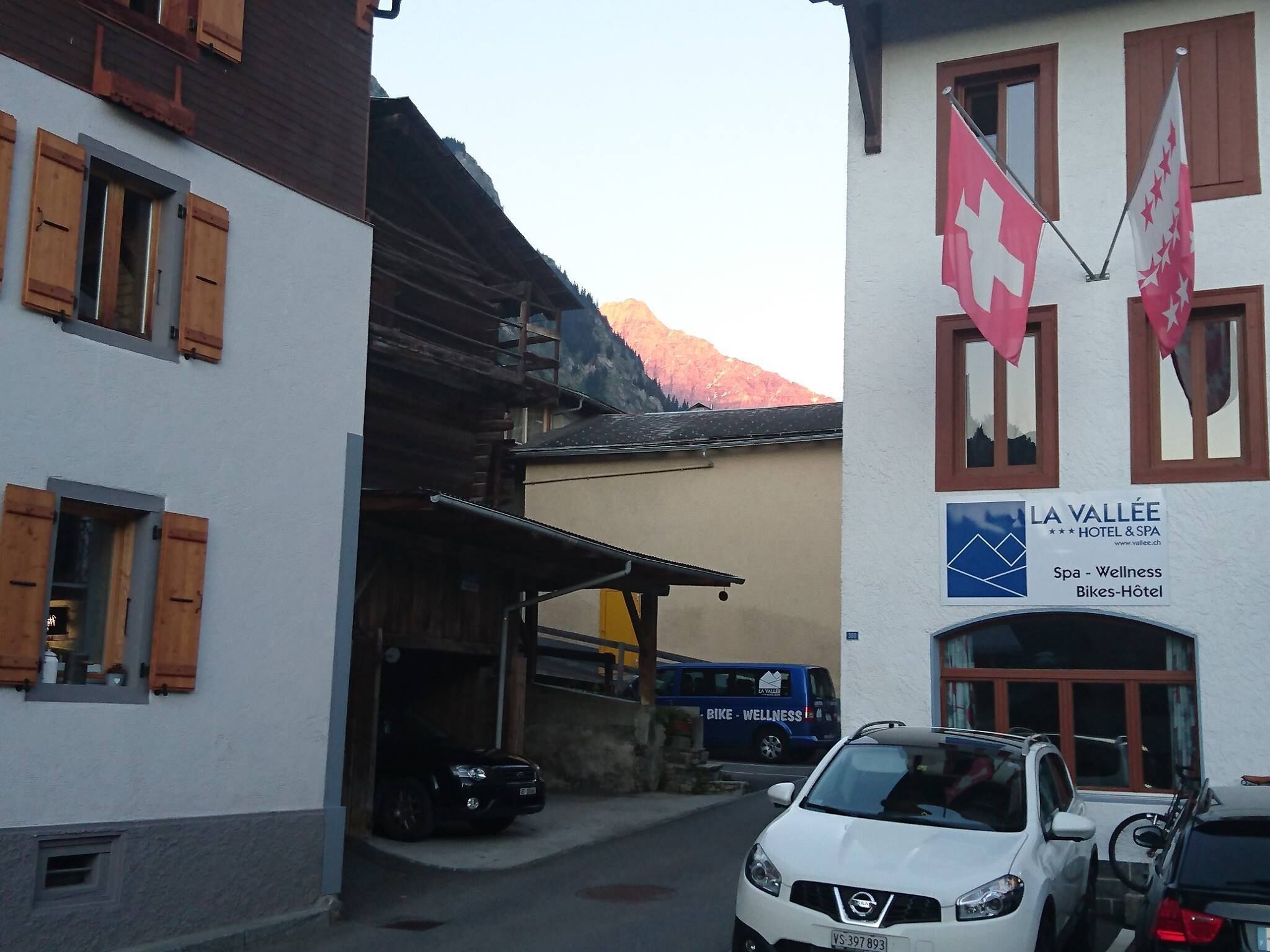 Une rue étroite bordée d’immeubles, avec une voiture et une camionnette garées devant un hôtel appelé « LA VALLÉE ».