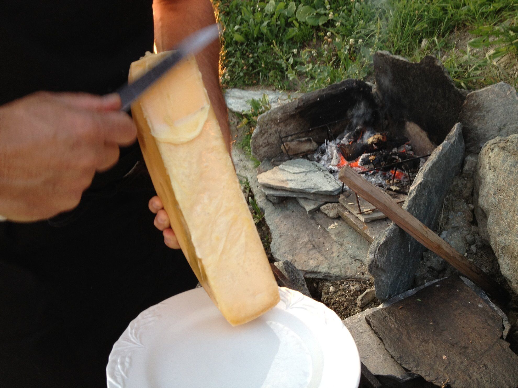 A person holding a long, rectangular object over a white plate, with a fire and rocks in the background.