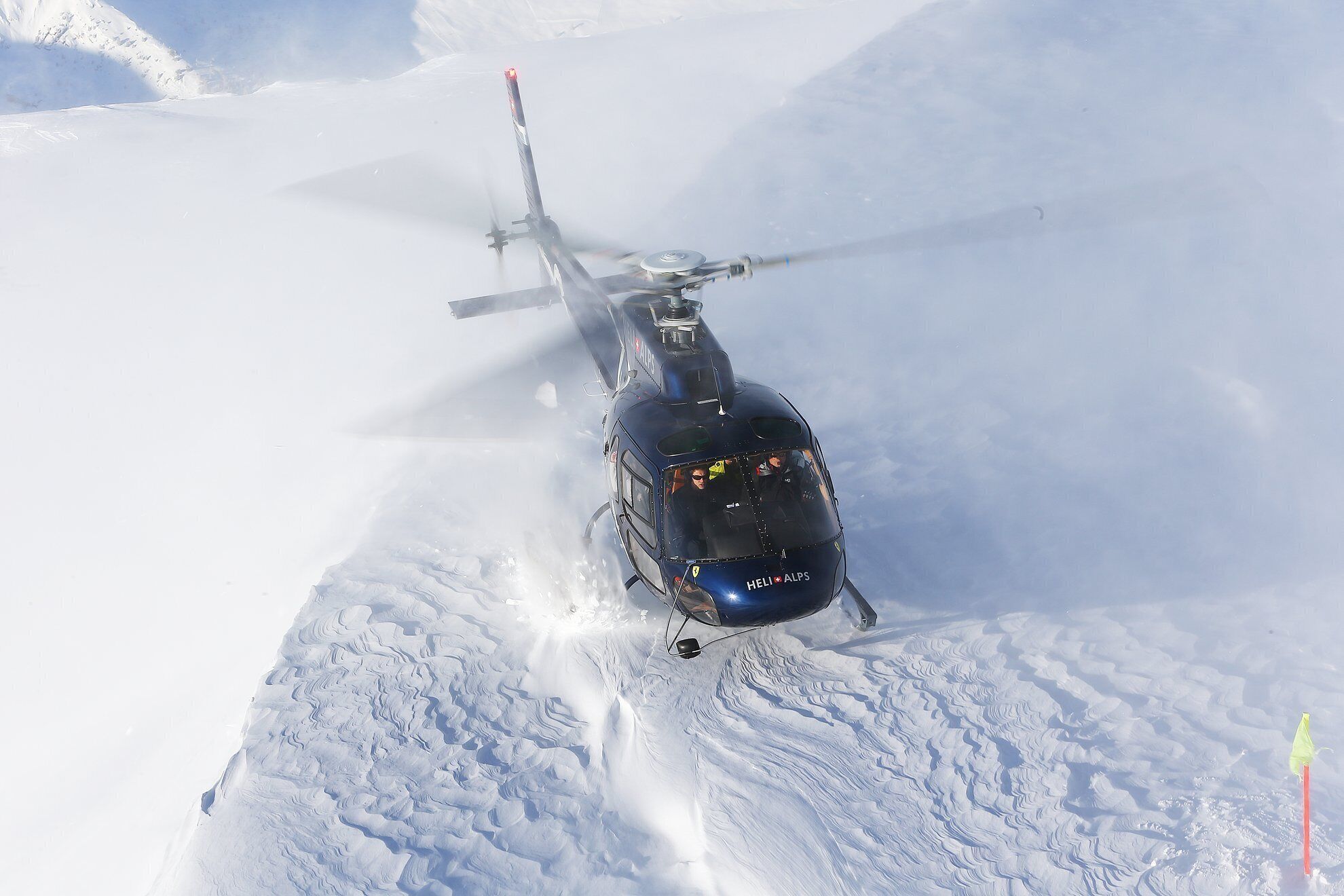A helicopter with "HELI ALPS" written on the side flying over snow-covered terrain.