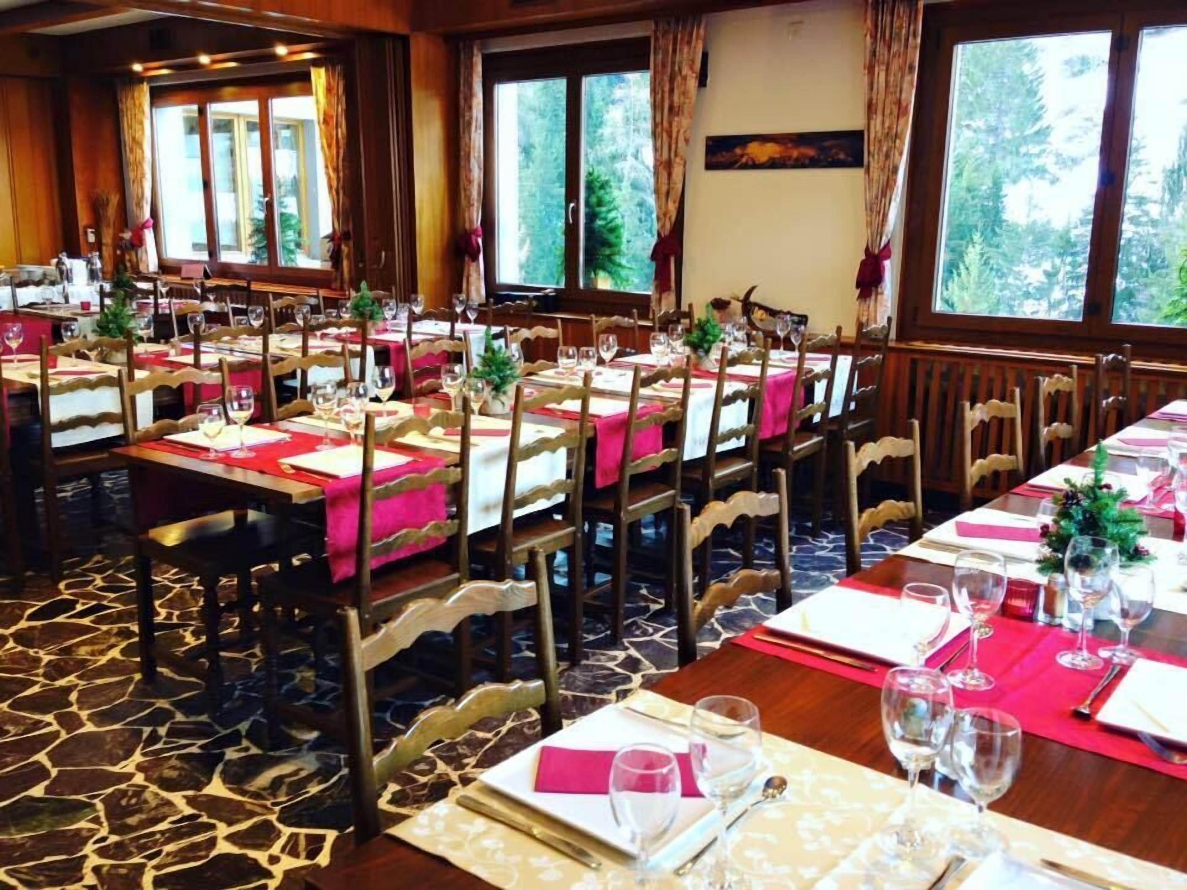 A dining area with multiple tables set for a meal, wooden chairs, and large windows offering a view of trees.