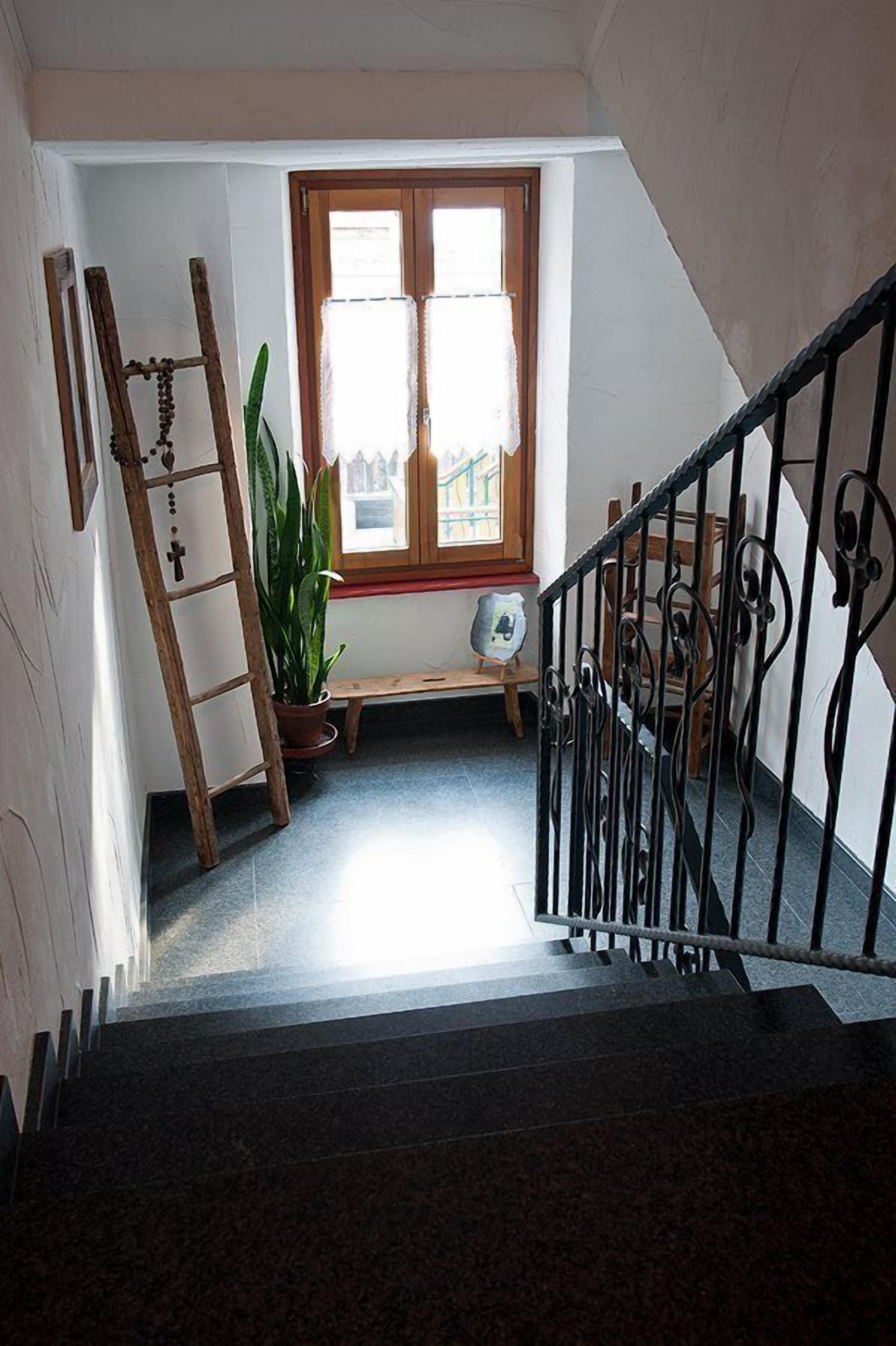 Una escalera con barandilla de madera y baranda de metal negro, que conduce a una habitación con ventana, planta en maceta y un pequeño banco.