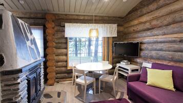 A log cabin interior with a stone fireplace, a round dining table with chairs, a purple sofa with a yellow pillow, and a mounted flat-screen TV.