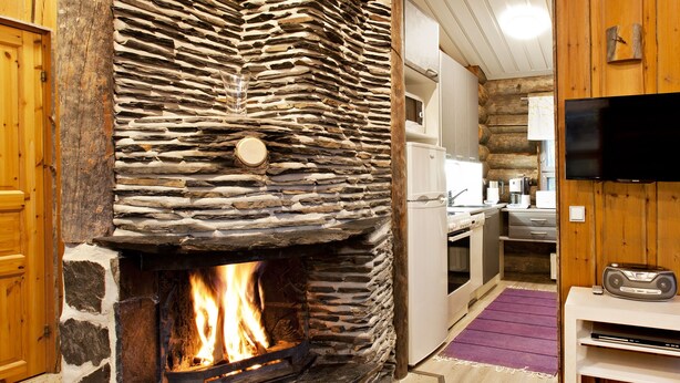 A stone fireplace with a fire, a wooden door, a kitchen area with appliances, and a television on a wooden wall. - Holiday Club Pyhäniemi (Kihnio)