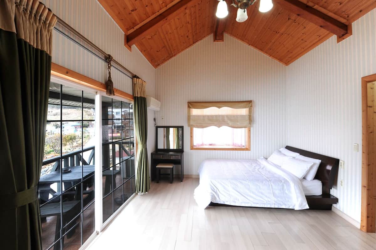 Un dormitorio con una cama grande, una ventana con persianas, techo de madera y un balcón con mesa y sillas.