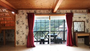 Une pièce avec des poutres apparentes, une grande fenêtre ornée de rideaux rouges, une table en bois et un miroir posé sur une commode.