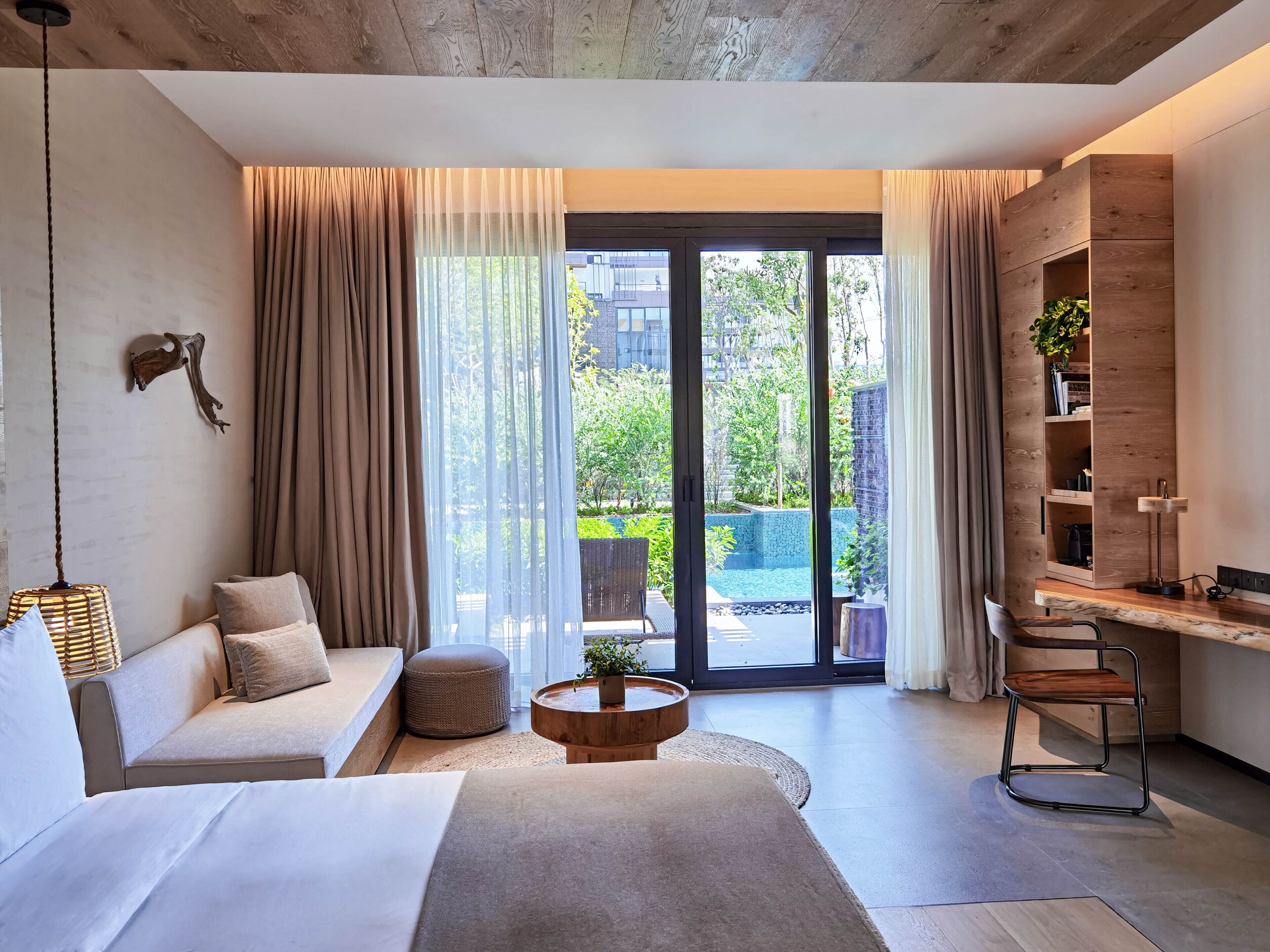 Habitación de hotel moderna con un amplio ventanal corredero que da a una zona de piscina, un sofá, una mesa de centro y un escritorio con una silla.