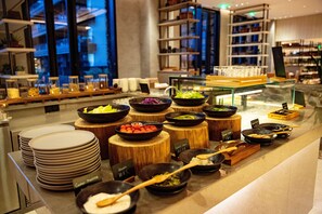 A buffet area with various dishes displayed on wooden stands, surrounded by stacks of plates and bowls. - Sunshine Yi Hotel Haitang Bay, Sanya (Sanya)