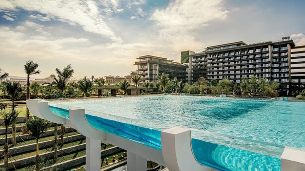 Ein großer, modern gestalteter Pool, umgeben von Palmen und mit Blick auf ein Hotel im Hintergrund.