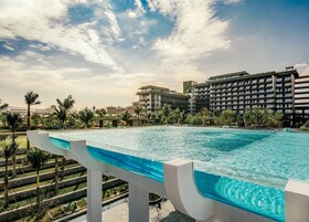 A large swimming pool with a modern design, surrounded by palm trees and a view of a hotel building in the background.