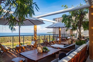 A terrace with wooden furniture, white umbrellas, and a view of the ocean.