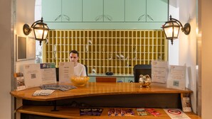 A reception desk with a man standing behind it, a yellow wall with shelves, and various informational brochures and signs. - MP Hostel Budapest (Budapest)