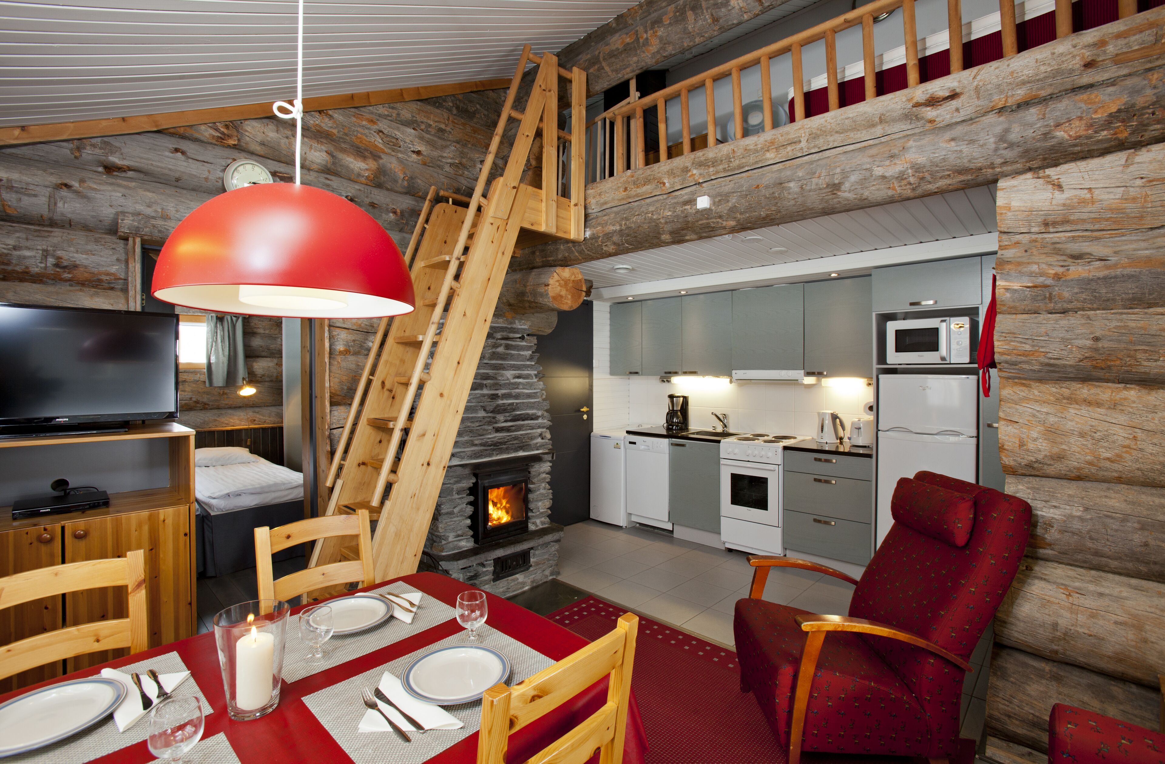Un intérieur de chalet chaleureux avec une cheminée en pierre, une table à manger en bois pour deux personnes, une cuisine équipée d’appareils modernes et une chambre à l’étage accessible par une échelle en bois.