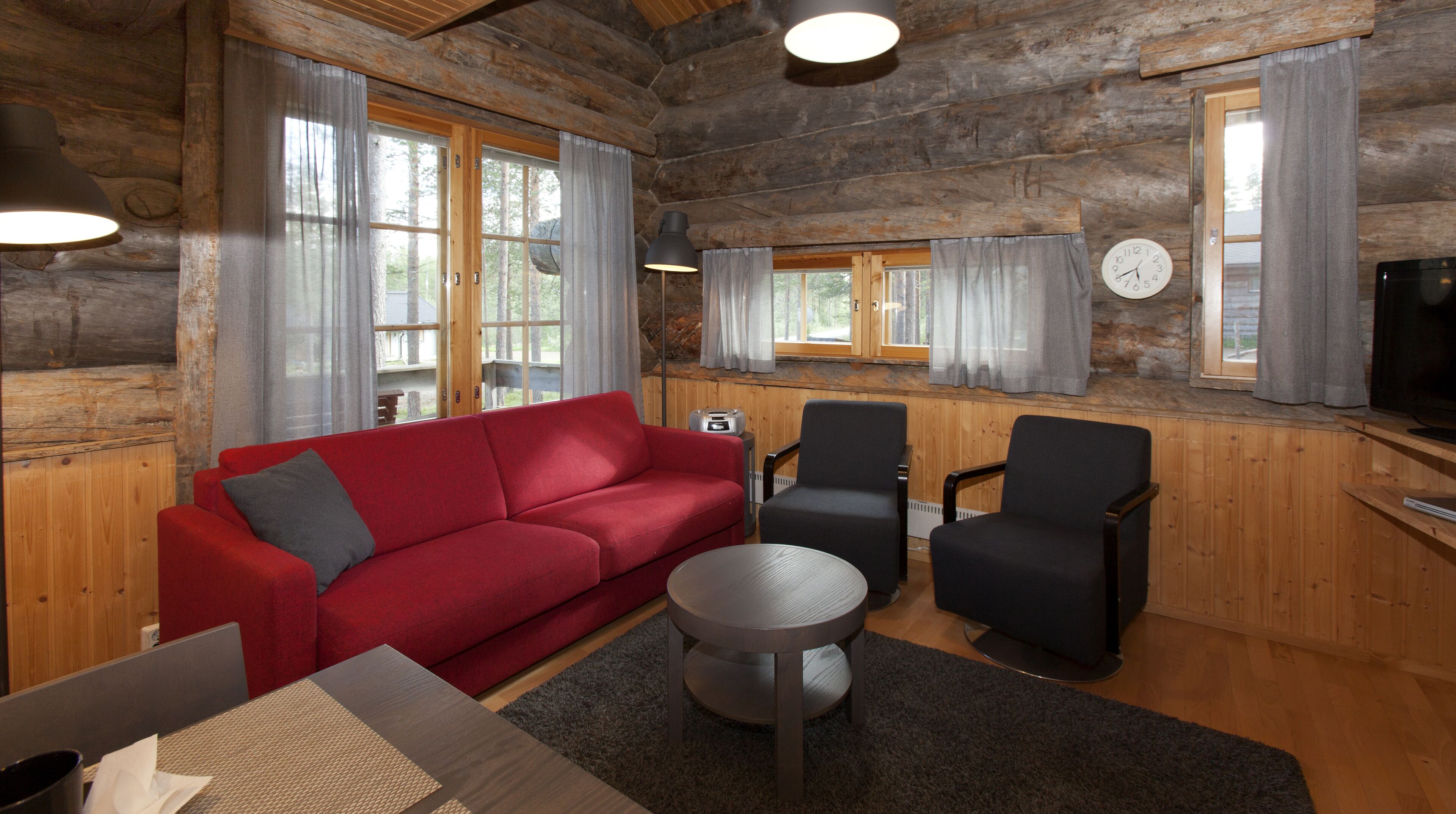 A log cabin interior with a red sofa, two black armchairs, a small round table, and a television.