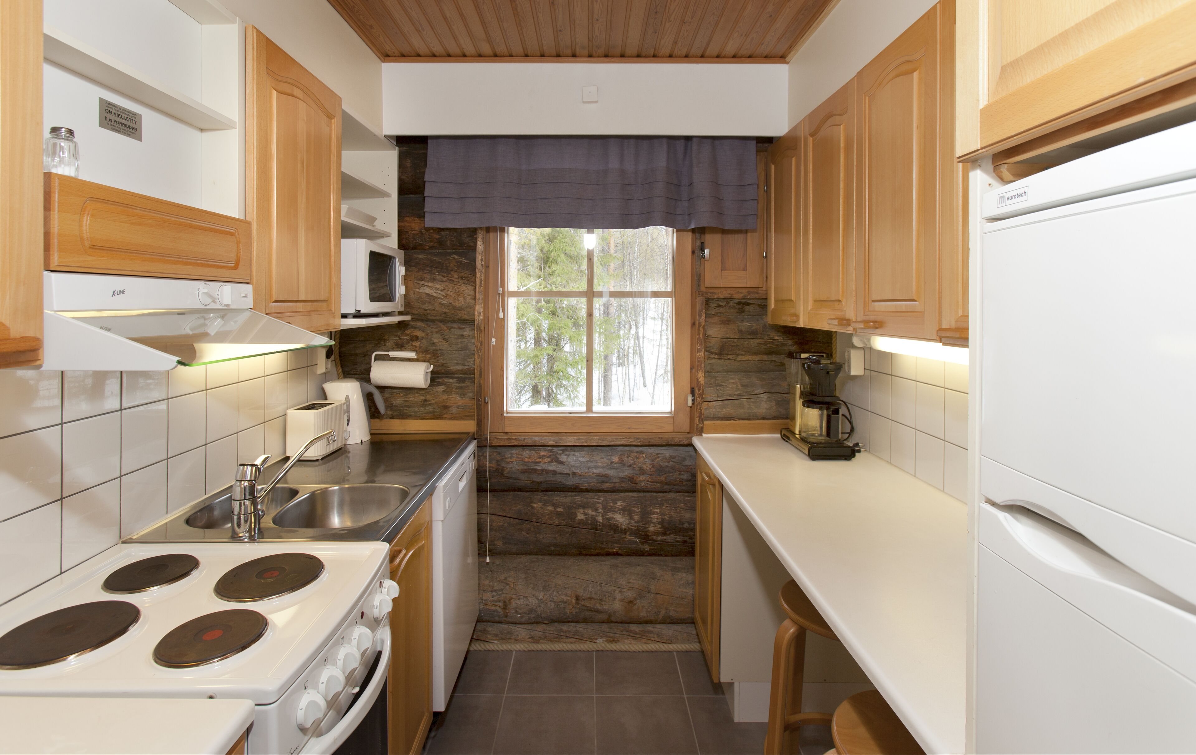 A compact kitchen with wooden cabinets, a stove, a microwave, and a window with a view of trees.