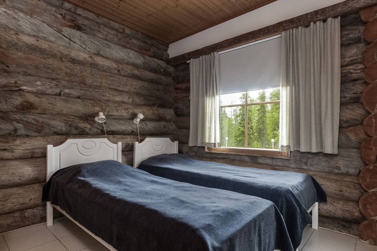 A log cabin room with two beds, a window with curtains, and a lamp on the wall.