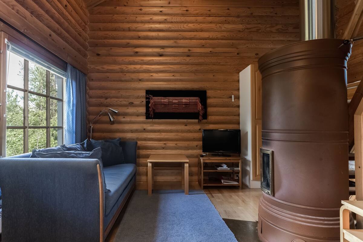 A cozy cabin interior with a wooden ceiling, a sofa, a television, and a large barrel stove.