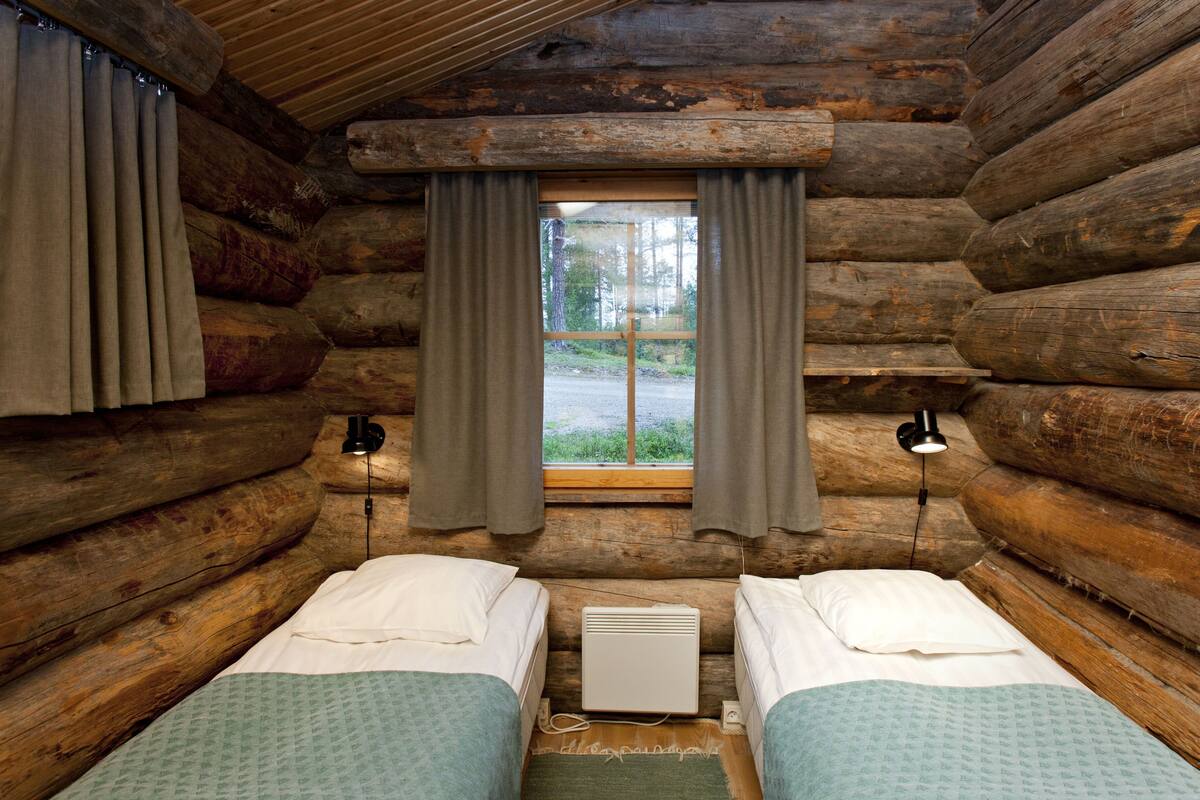 A log cabin room with two beds, a window with curtains, and a wall-mounted heater.