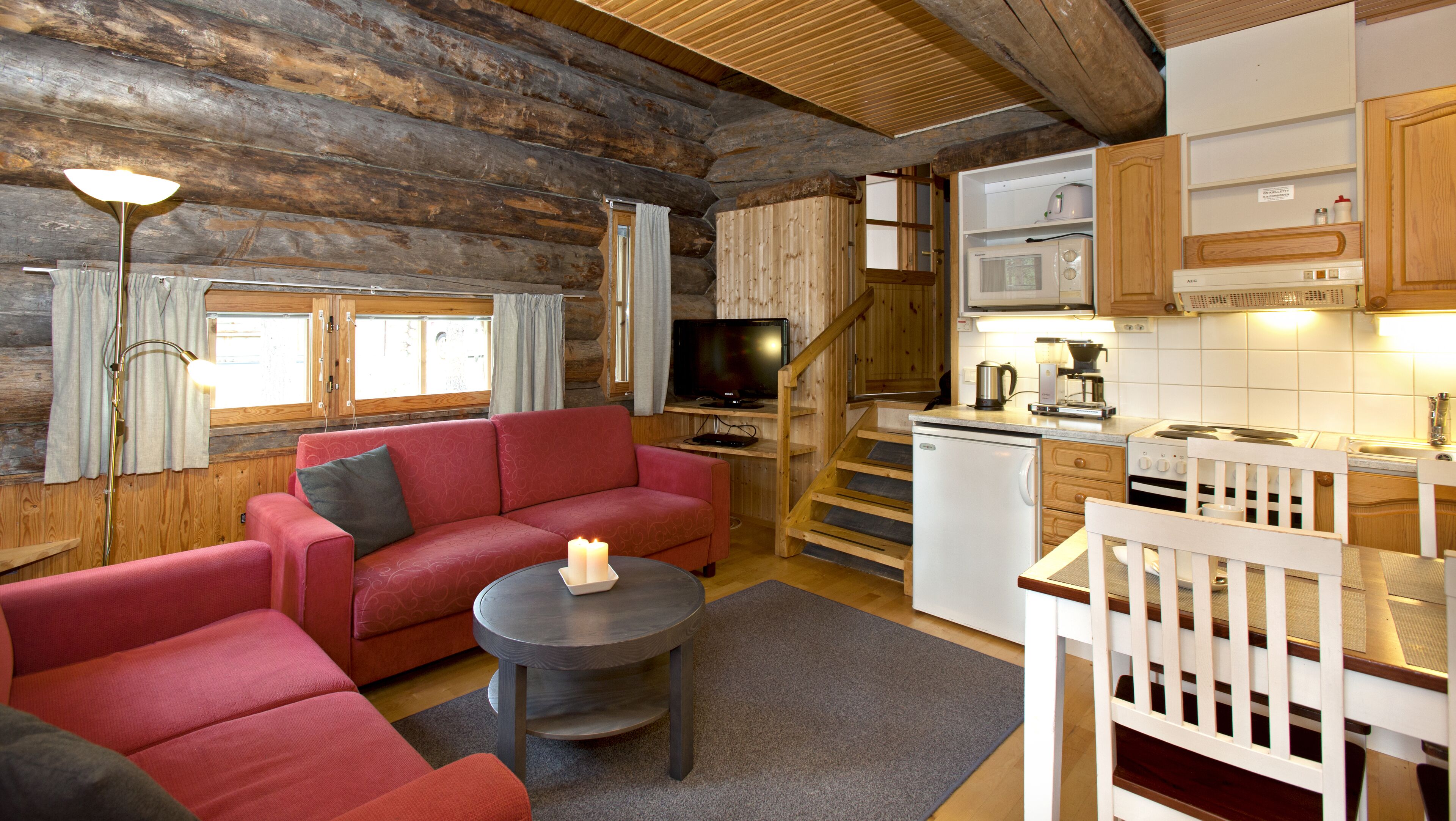 A cozy log cabin interior with a living area featuring a red sofa, a small round table with candles, and a television. A kitchen with wooden cabinets and a stove is visible in the background.