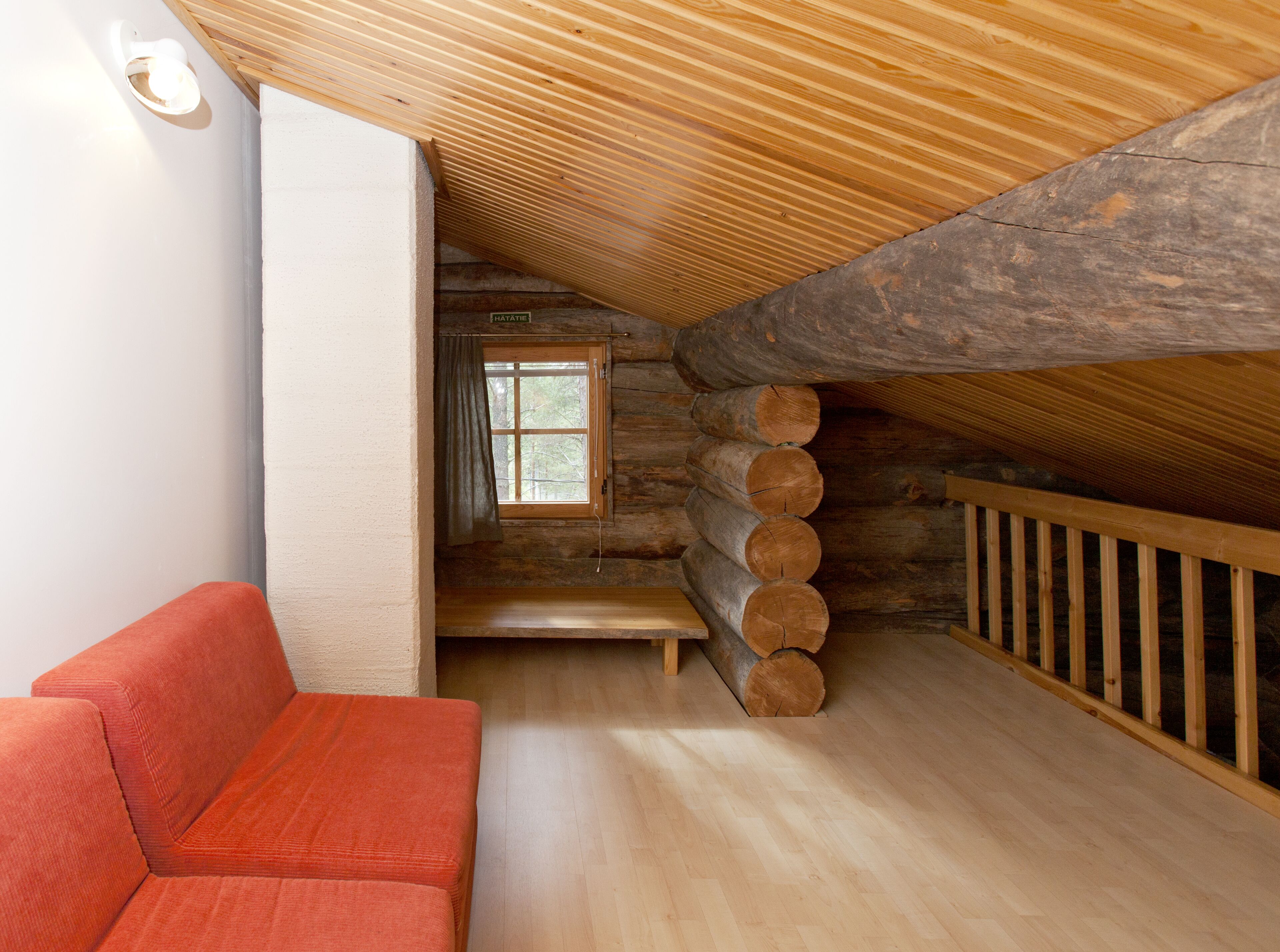 A log cabin interior with a wooden ceiling, a window with curtains, a wooden bench, and a red sofa.