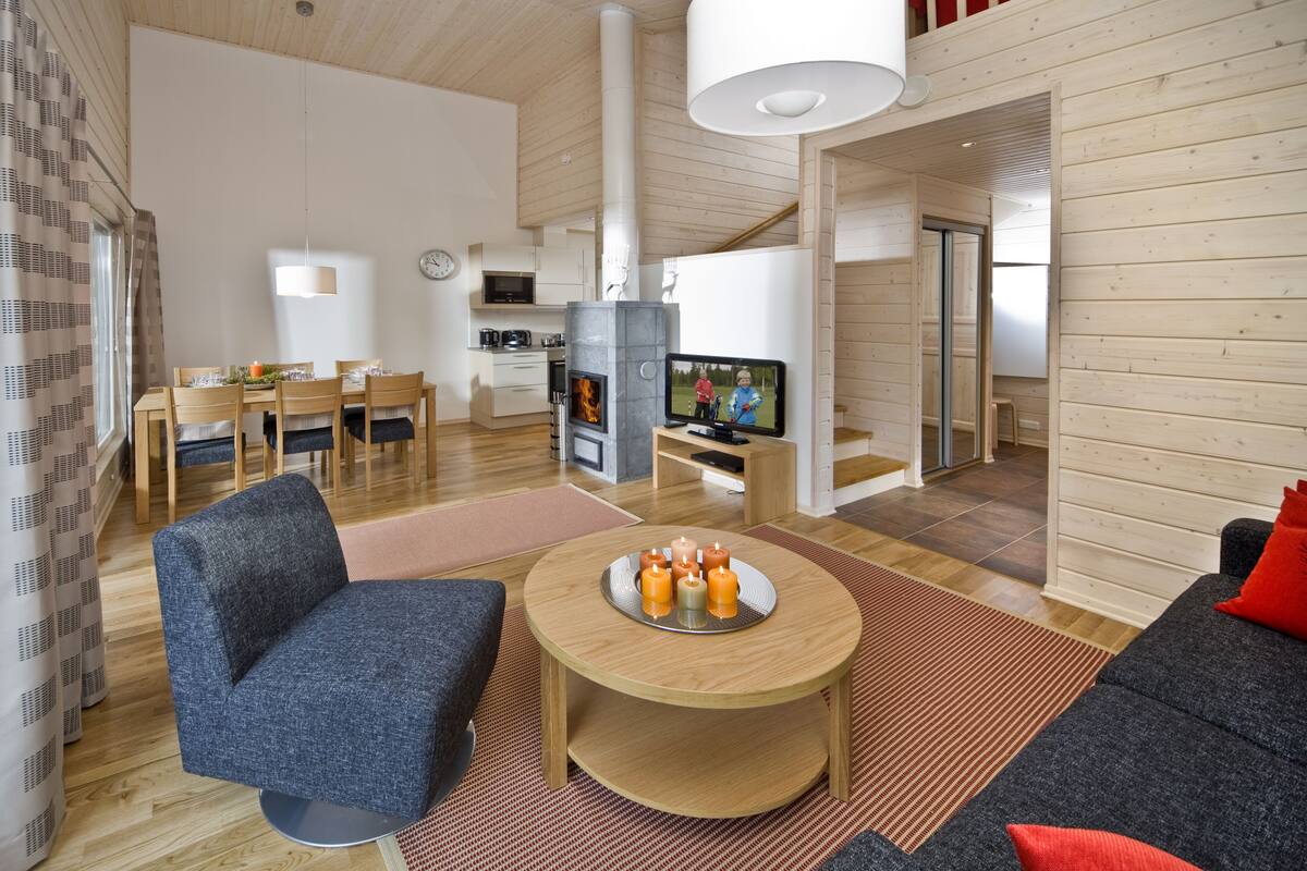A modern living room with a sofa, a coffee table, and a TV. There is a fireplace, a dining area, and a kitchen in the background.