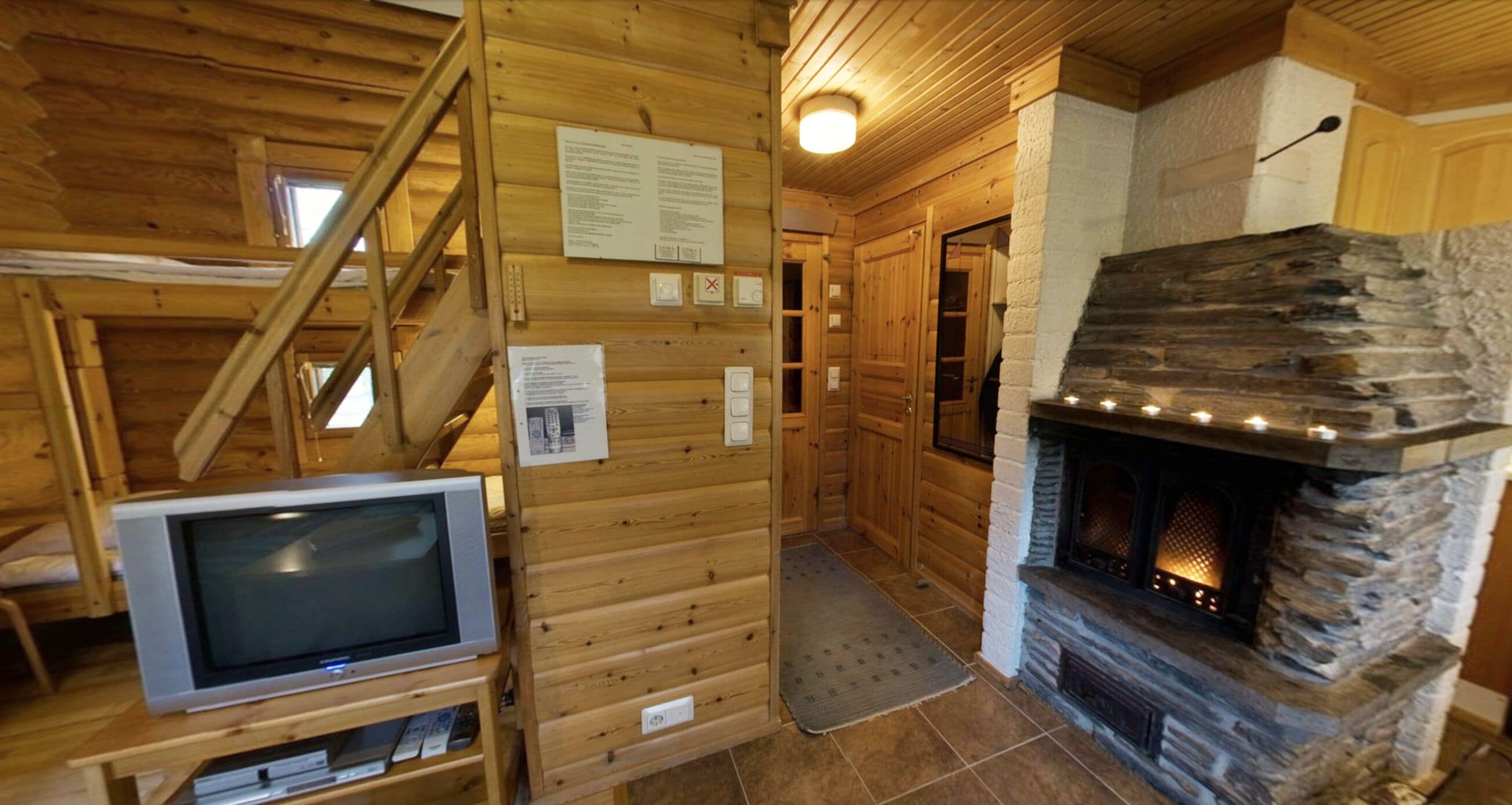 Interior de una cabaña de madera con chimenea de piedra, paredes de madera, televisor sobre un mueble bajo y una escalera.